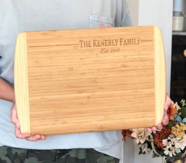 Two tone rectangle wood cutting board featuring two curved ends that are a lighter wood color than the center. The top right is engraved with a family name and date.