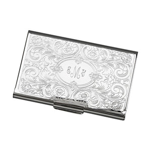 Feminine shiny silver metal business card holder engraved in a center oval with a monogram. Outside the oval shape is a paisley and scroll design embossed throughout.