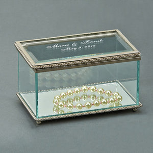 Rectangle shaped glass jewelry box with a silver braided edge. The top is engraved with with names and a wedding date in a frosty white color. The bottom of the jewelry box is a glass mirror.