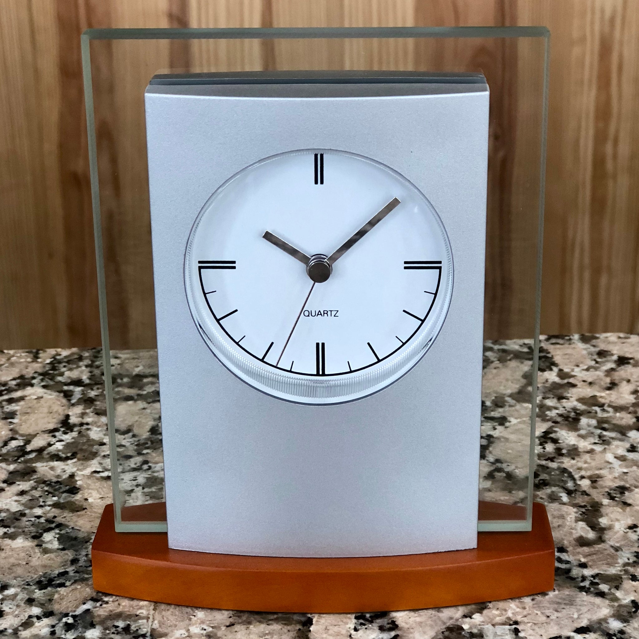 Silver and glass clock with light wood base featuring a white face with silver hands.