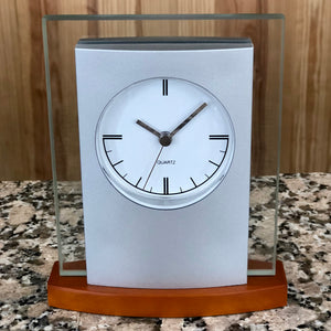 Silver and glass clock with light wood base featuring a white face with silver hands.