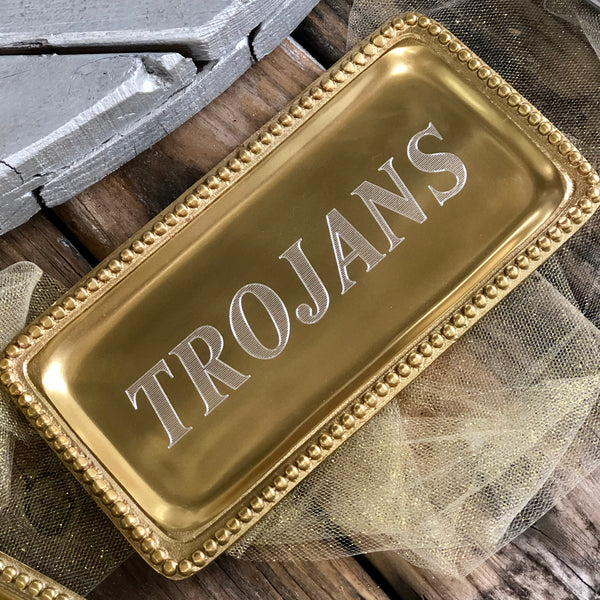 Rectangle shaped shiny gold tray with a beaded edge. The shiny gold tray is engraved to read "TROJANS" in the center.