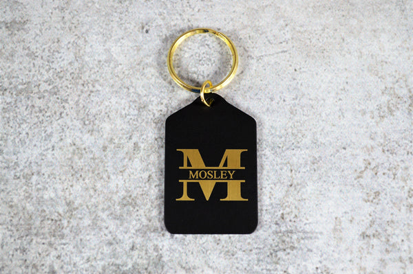 Rectangle shaped black brass keychain with peaked top and shiny gold key ring. The keychain is engraved with a large "M" and a last name going through the center of it. Engraving is brass colored.