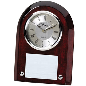Small high gloss rosewood clock with a curved top and beveled edges. Features a silver face with black roman numerals and  an engraved plate below the face.